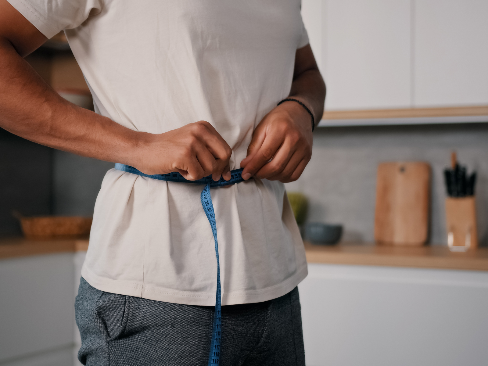Un hombre se mide la cintura con una cinta métrica azul en una cocina, como imagen de la pérdida de peso y el control del apetito; descubra cómo la tirzepatida reduce el apetito.