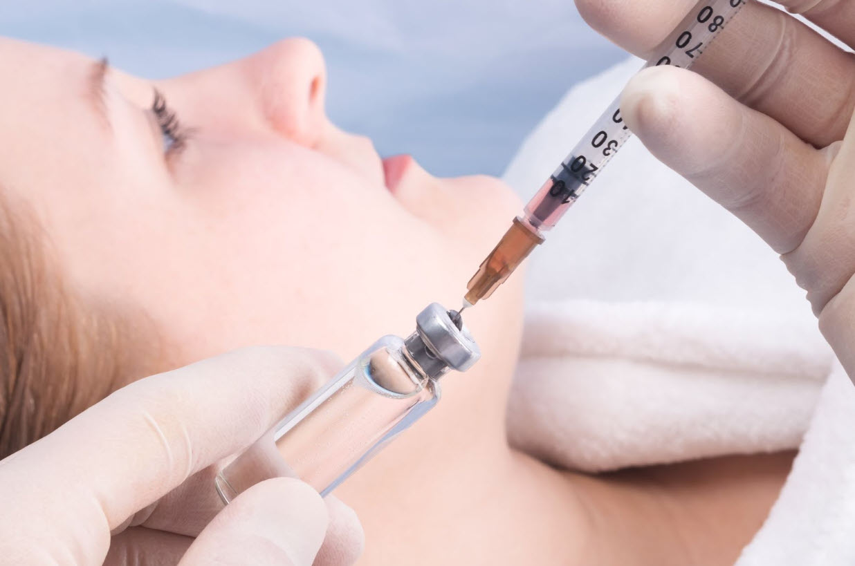 Un profesional de la salud con guantes prepara una jeringa extrayendo líquido de un vial, con un paciente recostado en un entorno clínico, lo que ilustra un procedimiento médico seguro y controlado y plantea la pregunta: ¿es segura la terapia con péptidos para distintos tratamientos de salud y bienestar?