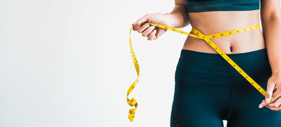 Una mujer en forma, con ropa deportiva oscura, se pasa una cinta métrica amarilla alrededor de la cintura: una imagen de pérdida de peso y transformación corporal que destaca los beneficios de la tesamorelina, que ayuda a reducir la grasa abdominal, mejorar el metabolismo y apoyar el bienestar general.