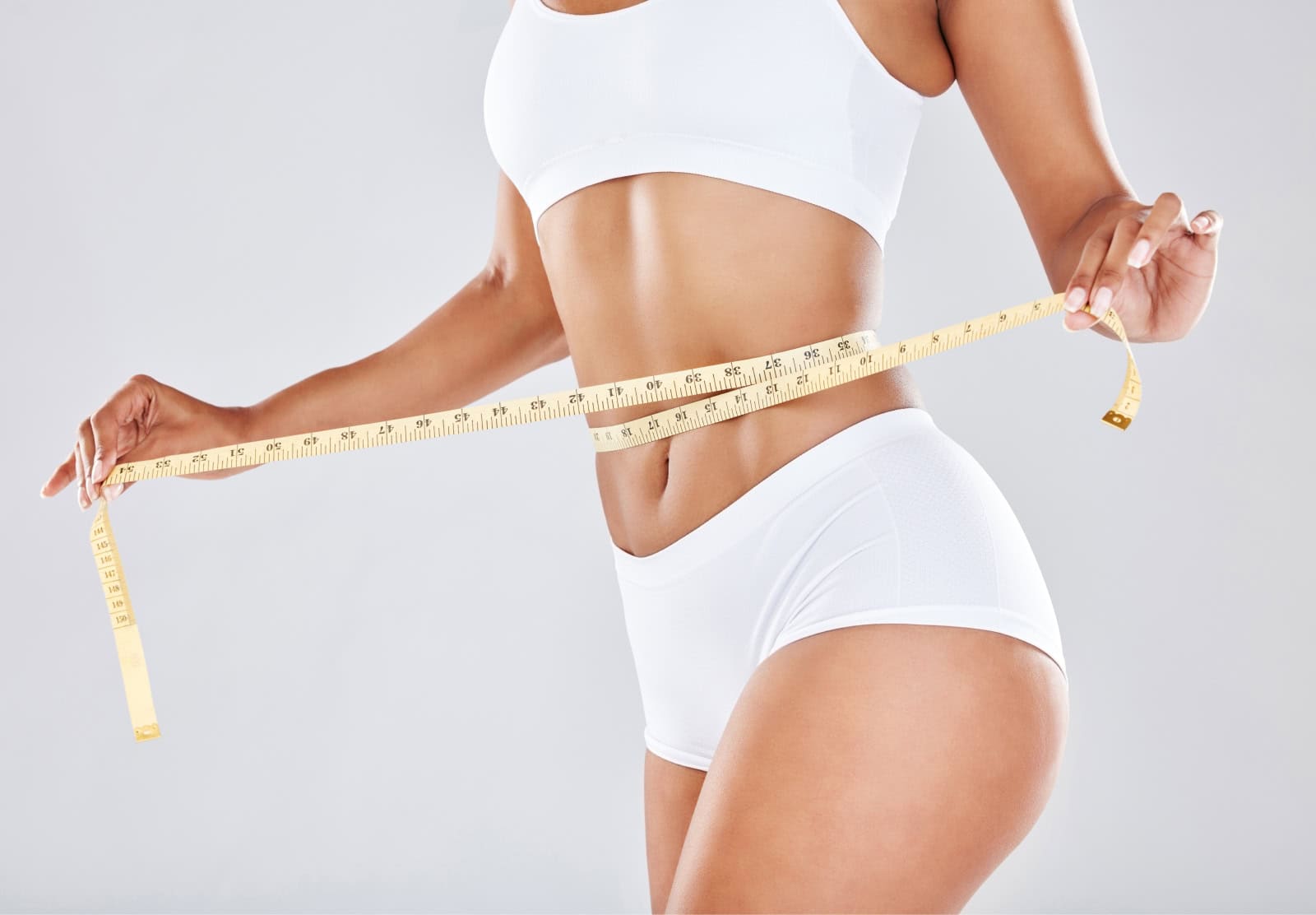 Primer plano del abdomen de una mujer con ropa deportiva blanca que se pasa una cinta métrica alrededor de la cintura, símbolo de los resultados de los programas de pérdida de peso médica, que favorecen un manejo saludable del peso y la confianza en el propio cuerpo.